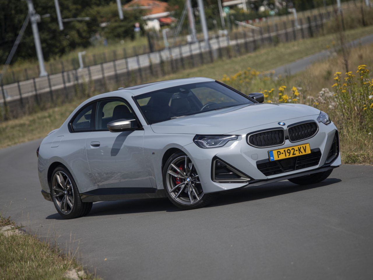Een slanke zilveren BMW M240i met kenteken "P-192-KV" rijdt op een bochtige weg omgeven door gras en wilde bloemen.