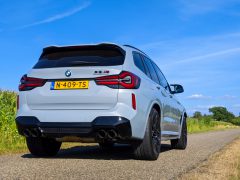 Een achteraanzicht van een grijze BMW X3 M met een geel kenteken geparkeerd op een smalle, verharde weg. Het omringende groene gras en de helderblauwe lucht versterken het strakke design van de BMW X3 M.