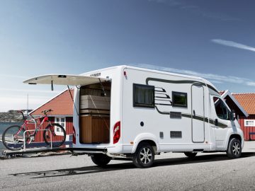 Een witte camper staat geparkeerd op een verhard oppervlak met de achterdeur open, waardoor een binnencompartiment en een gemonteerde fiets zichtbaar worden. Op de achtergrond zijn kleine gebouwen met rode daken zichtbaar.