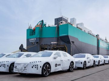 Meerdere auto's, waaronder verschillende NIO ET7-modellen, bedekt met beschermend wit materiaal, staan opgesteld op een kade bij een groot vrachtschip met de naam "Carmen" op de boeg.