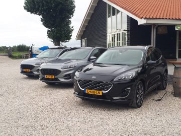 Drie zwarte SUV's, waaronder een slanke plug-in hybride, staan geparkeerd op een grindoprit voor een gebouw met een rood pannendak.