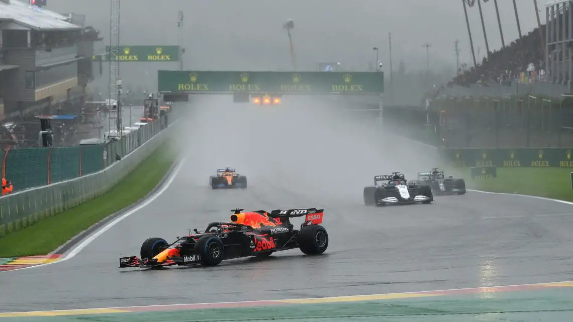 Verschillende Formule 1-auto's racen op een natte baan tijdens een regenachtig evenement in Spa. De leidende auto wordt op de voet gevolgd door anderen, met spatten water uit de wielen. Hoe laat begint de kwalificatie van Spa?