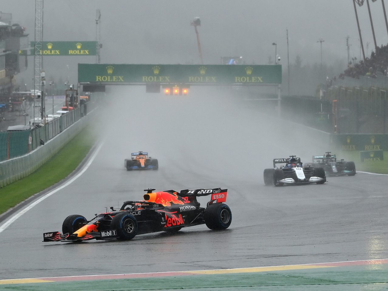 Verschillende Formule 1-auto's racen op een natte baan tijdens een regenachtig evenement in Spa. De leidende auto wordt op de voet gevolgd door anderen, met spatten water uit de wielen. Hoe laat begint de kwalificatie van Spa?