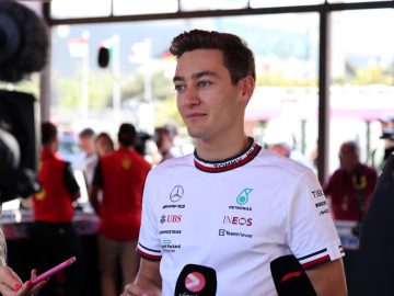 Een man in een wit sportshirt met diverse sponsorlogo's staat de media te woord terwijl meerdere microfoons op hem gericht zijn. George Russell wordt omringd door mensen en vlaggen die op de achtergrond zichtbaar zijn.