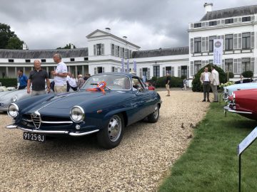 Een klassieke blauwe auto wordt tentoongesteld op een onverharde oprit tijdens een Concours d'Elegance-evenement buiten, voor een wit gebouw. Verschillende mensen observeren de auto.