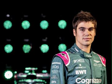 Lance Stroll, gekleed in een racepak, staat zelfverzekerd voor ronde lichten, met zijn gestroomlijnde auto gedeeltelijk zichtbaar op de achtergrond.