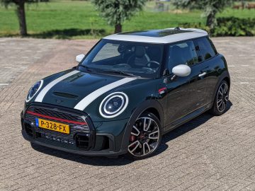 Op een verhard terrein met gras en bomen op de achtergrond staat een donkergroene Mini John Cooper Works met witte racestrepen en een Nederlands kenteken geparkeerd.