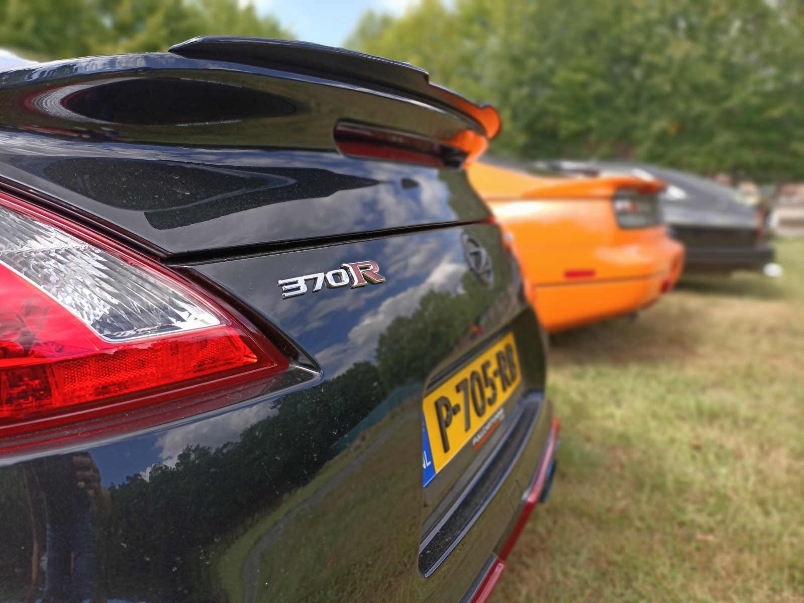 Close-up van de achterkant van een zwarte Nissan Z 370R-sportwagen met zichtbaar kenteken, geparkeerd naast een oranje auto en een ander voertuig in een grasveld. Bomen zijn op de achtergrond.