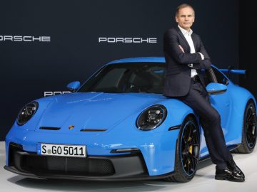 Een man in pak leunt tegen een blauwe Porsche-sportwagen, met 'Porsche'-logo's op de achtergrond. Deze Porsche, ontworpen om op e-brandstoffen te rijden, benadrukt de toekomst van het autorijden en is voorzien van een kentekenplaat met de tekst ‘S-GO 5011.