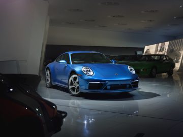 In een goed verlichte showroom wordt een blauwe Porsche 911 sportwagen tentoongesteld met andere auto's op de achtergrond.