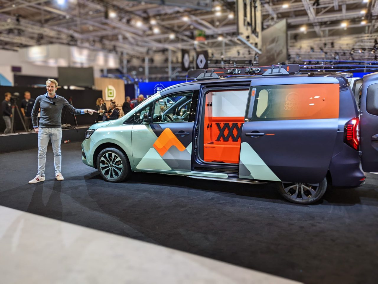 Een man staat naast een zwart-zilveren custom Renault-camper met oranje accenten die tijdens een indoorevenement wordt tentoongesteld en wijst ernaartoe. Het busje heeft schuifdeuren, waardoor een gedetailleerd interieur zichtbaar is.
