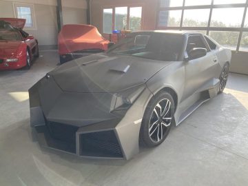 Een moderne grijze sportwagen met een hoekig ontwerp staat binnen geparkeerd naast een voertuig bedekt met een rode stof, waardoor het bijna een mysterieuze replica lijkt. De scherpe lijnen en zwarte velgen van de auto zijn duidelijk zichtbaar.