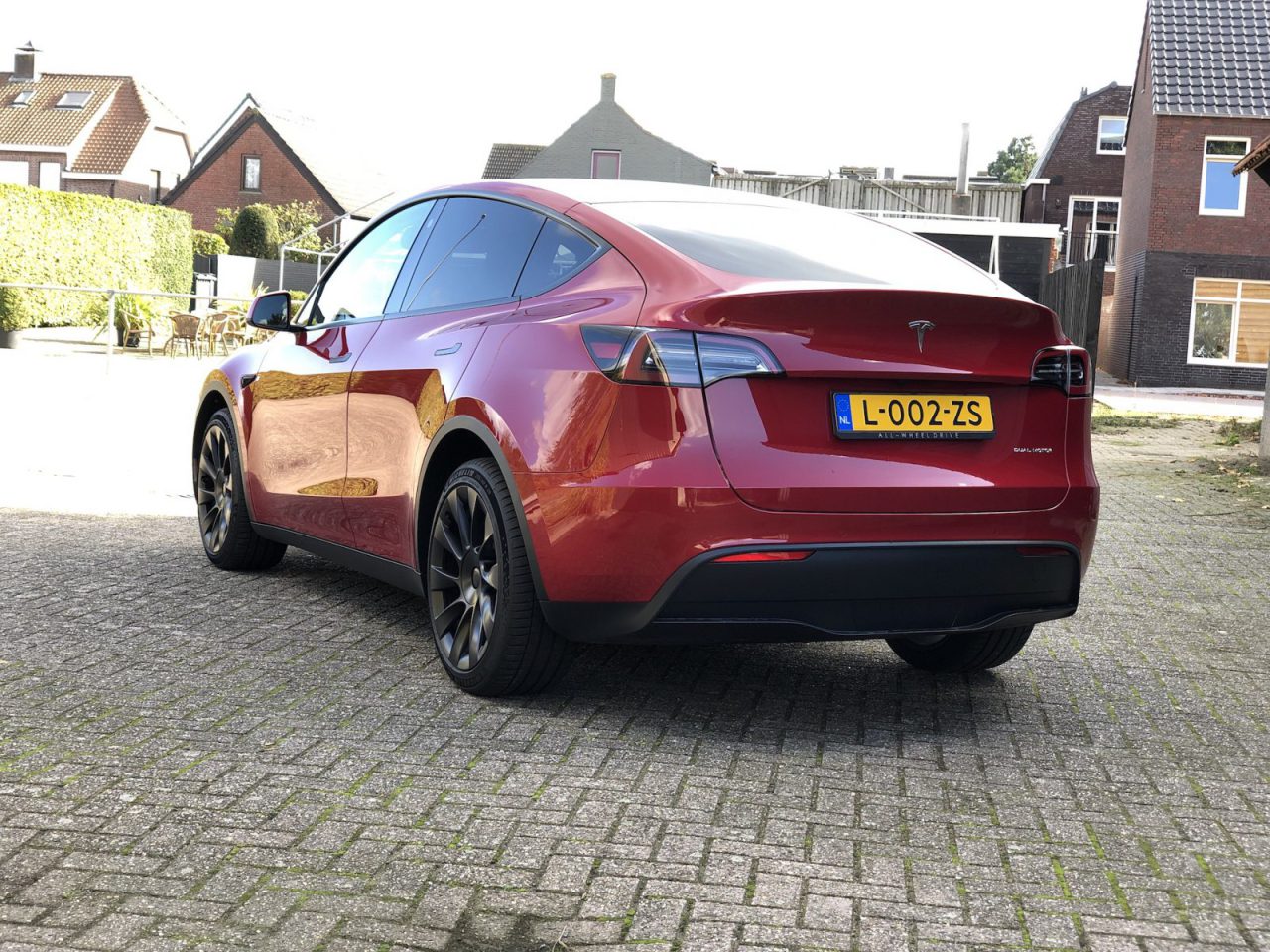 Een strakke rode Tesla staat geparkeerd in een geplaveide straat in een schilderachtige woonwijk, met charmante bakstenen huizen die een schilderachtige achtergrond vormen. Op het kenteken van de auto staat "L-002-ZS.