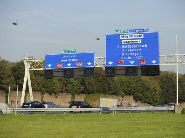 Een snelwegbord in Nederland geeft de routebeschrijving naar Arnhem, Amersfoort, Utrechtse Ring en Jaarbeurs aan. In de scène zijn enkele auto's te zien die bij heldere hemel op een meerbaansweg rijden, waarbij langs de berm enkele wegwerkzaamheden plaatsvinden.
