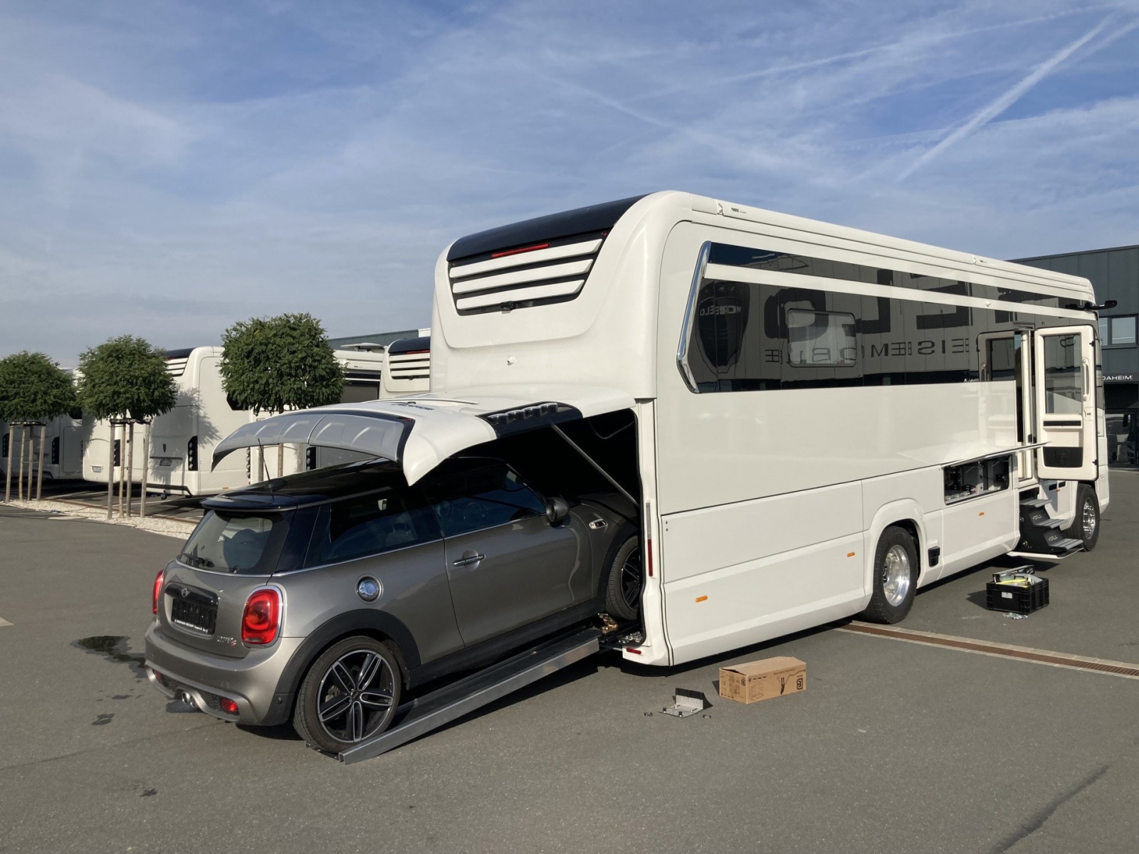 Een kleine auto wordt via een hydraulische oprit achterin een duurste camper geladen, met op de achtergrond bomen en gebouwen zichtbaar.