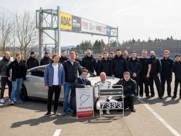 Een groep mensen staat rond een auto op het Nürburgring-circuit, met een bord met de tekst "7:33", met op de achtergrond verschillende spandoeken en logo's zichtbaar.