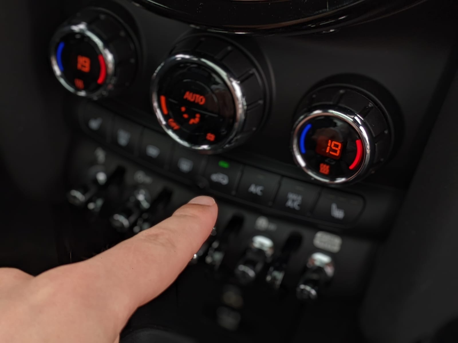 Een hand past de klimaatinstellingen in een auto aan, met temperatuurdisplays die 19 graden Celsius aangeven en verschillende bedieningsknoppen zichtbaar, waaronder AC en stoelverwarming: een perfecte zomertip om brandstof te besparen en tegelijkertijd comfortabel te blijven.