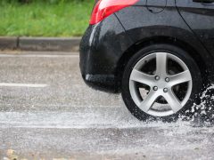 Close-up van de achterband van een zwarte auto die door een plas rijdt op een natte, gladde weg, waardoor water spettert. De achtergrond toont een met gras begroeide kant van de weg.
