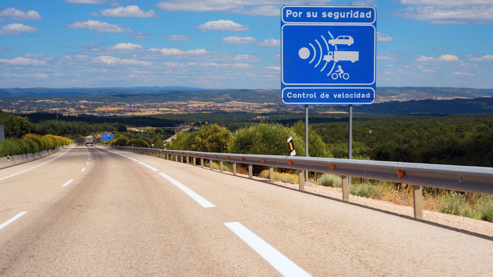 In de verte strekt zich een snelweg uit met een blauw bord met de tekst "Por su seguridad, Control de velocidad" dat snelheidscontrole voor de veiligheid aangeeft. Het heuvelachtige landschap met verspreide vegetatie ligt onder de zonnige hemel. Houd er rekening mee dat het negeren van dergelijke signalen kan leiden tot een boete in het buitenland.