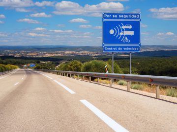 In de verte strekt zich een snelweg uit met een blauw bord met de tekst "Por su seguridad, Control de velocidad" dat snelheidscontrole voor de veiligheid aangeeft. Het heuvelachtige landschap met verspreide vegetatie ligt onder de zonnige hemel. Houd er rekening mee dat het negeren van dergelijke signalen kan leiden tot een boete in het buitenland.