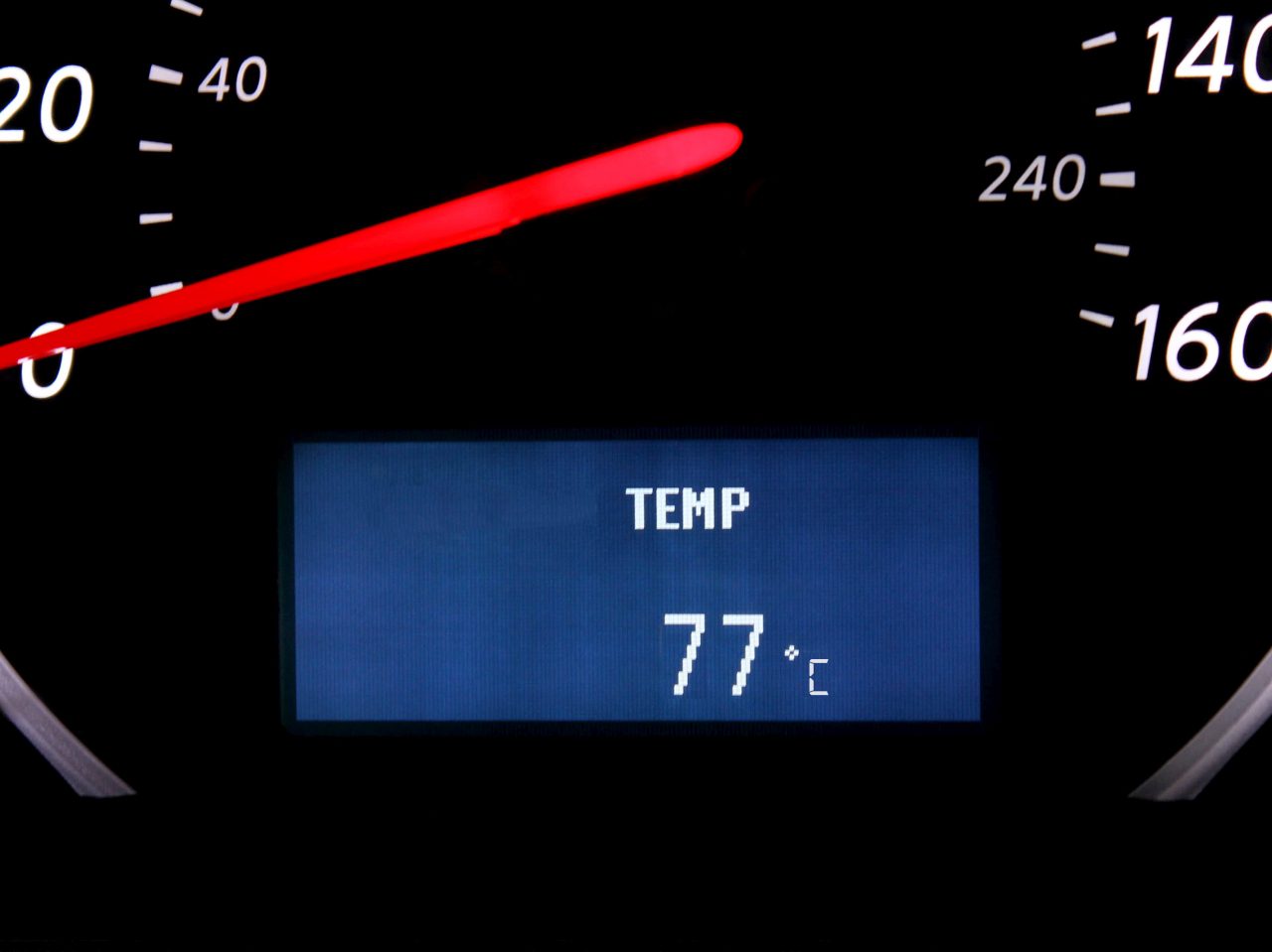 Een autotemperatuurmeter die 77 graden Celsius aangeeft, met een rode naald die tussen 0 en 20 op de snelheidsmeter wijst, wat de intense hitte van de motor van het voertuig benadrukt.