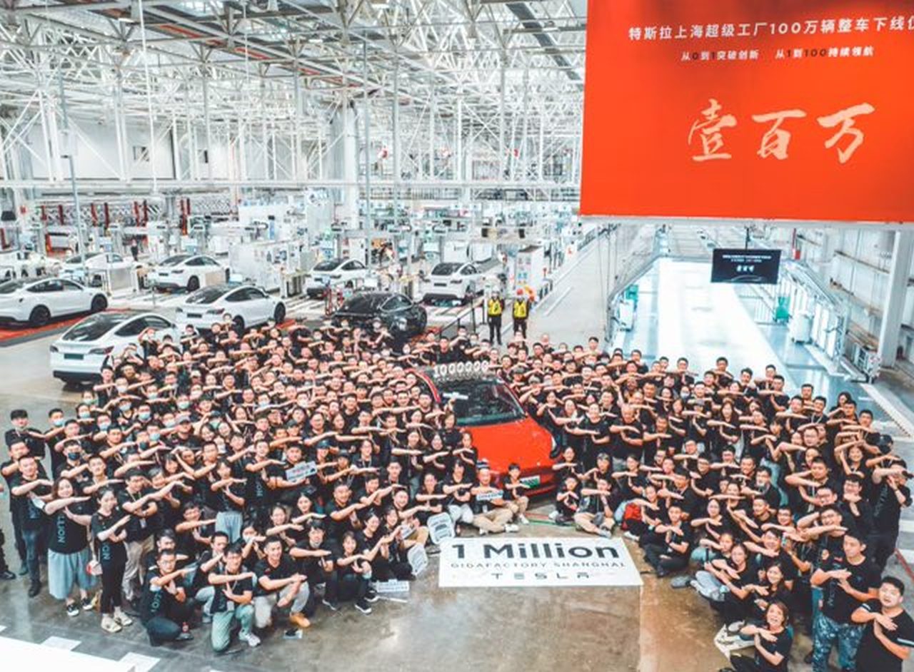 Een grote groep mensen in bijpassende zwarte shirts viert feest rond een rode Tesla in een fabrieksomgeving, met een spandoek met de tekst "1 Million" voor de auto geplaatst.