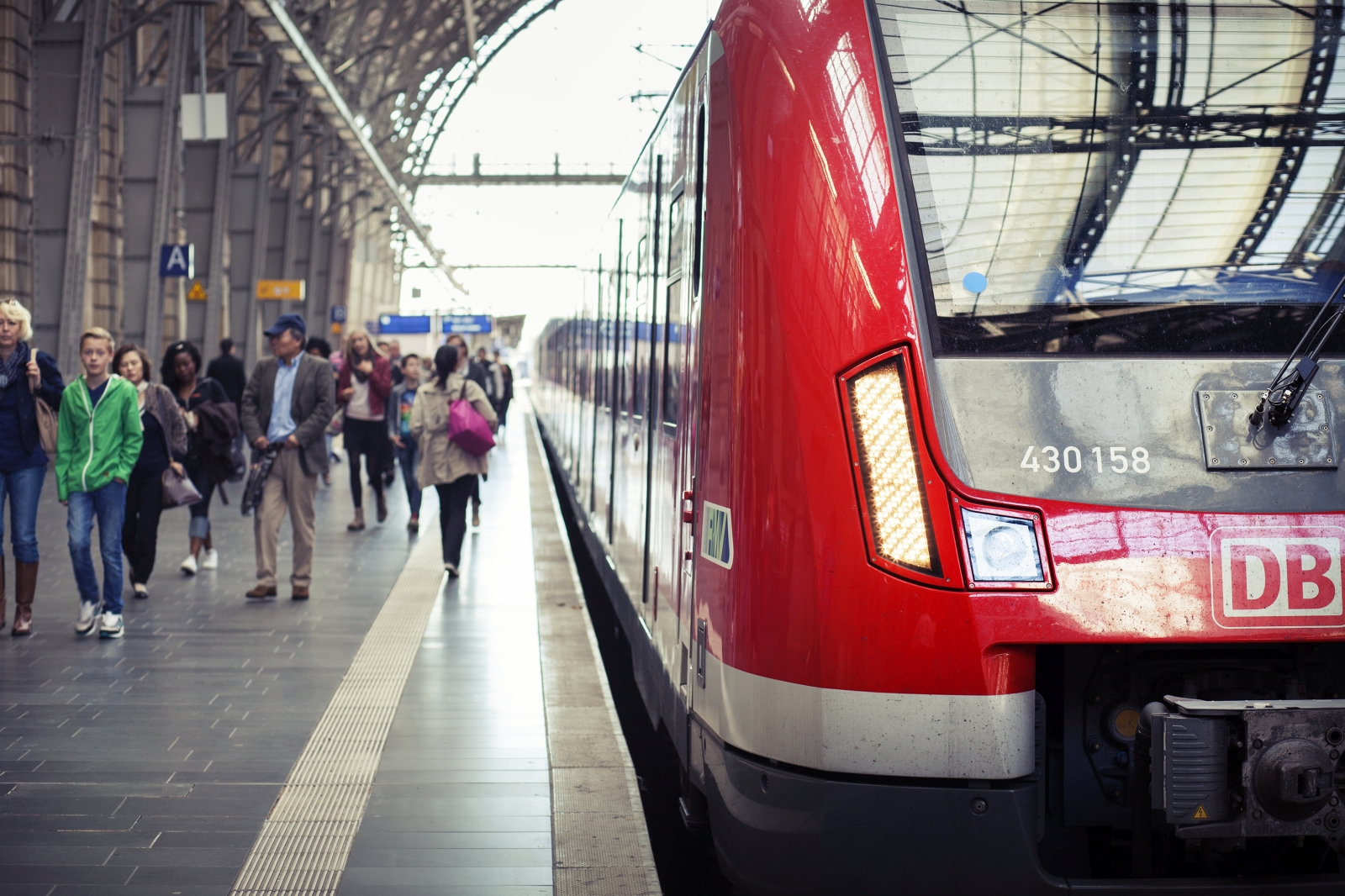 Op een perron van een groot treinstation staat een rode trein gestationeerd terwijl passagiers erlangs lopen, verlangend naar hun treinreisavontuur. Het nummer van de trein, 430 158, is zichtbaar op de voorkant.