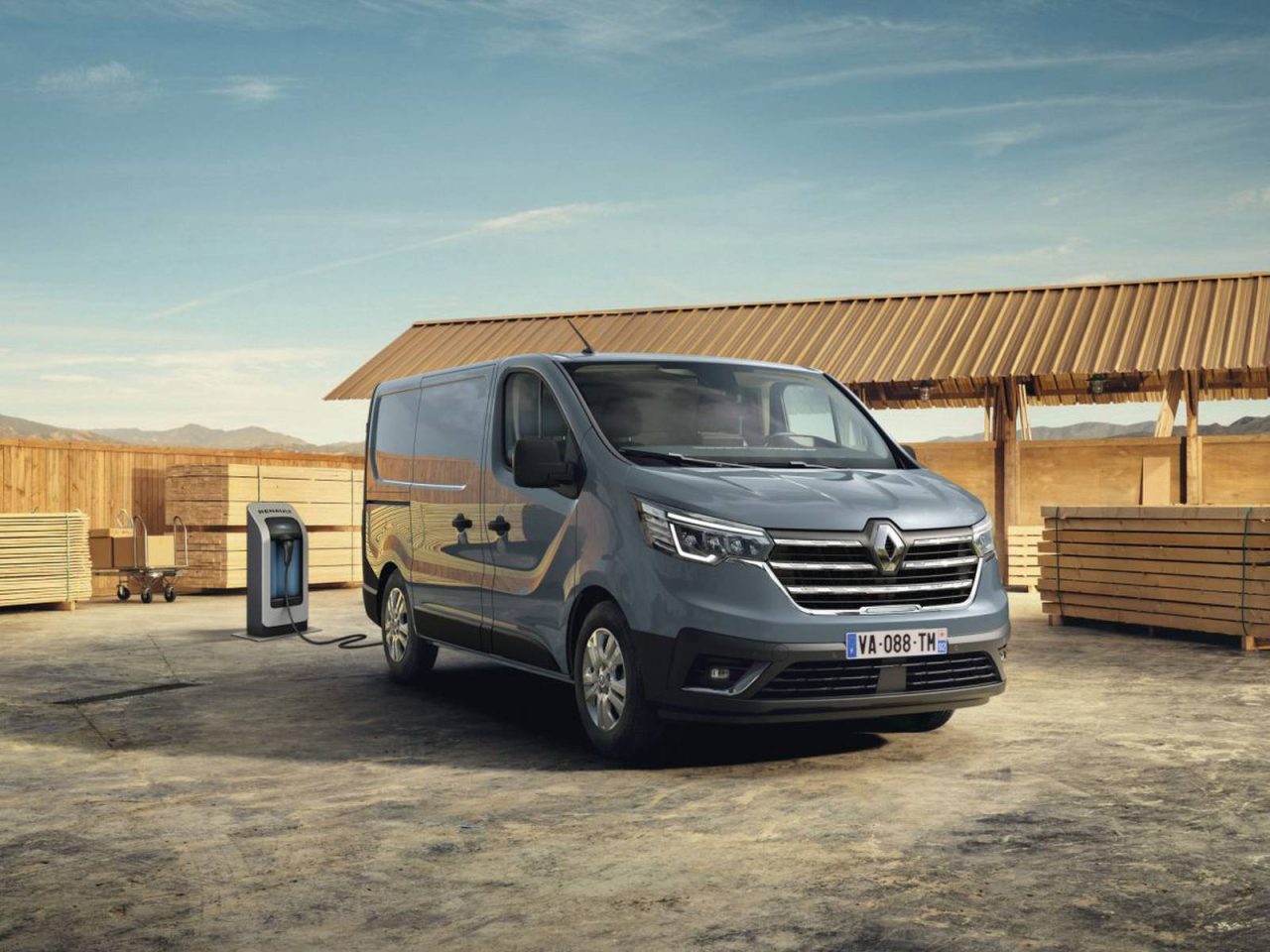 Een grijs Renault Trafic-busje staat geparkeerd bij een laadstation in een open ruimte met houten constructies op de achtergrond.