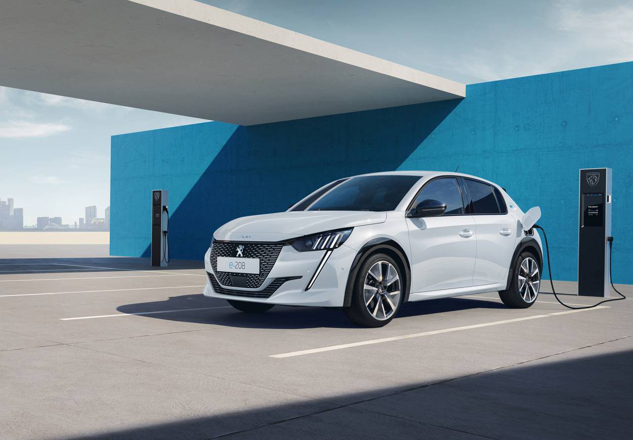 Een witte Peugeot e-208 staat geparkeerd bij een laadstation onder een betonnen overkapping met op de achtergrond de skyline van de stad. De auto wordt aangesloten op het laadstation.