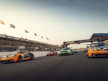 Verschillende Porsche-raceauto's, overwegend oranje en groen, racen onder een heldere hemel over een racecircuit, met toeschouwerstribunes en vlaggen zichtbaar op de achtergrond.