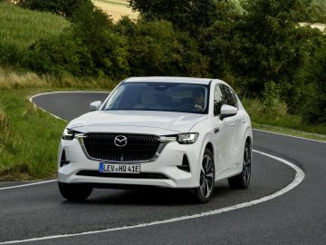 Een strakke witte Mazda CX-60 SUV navigeert moeiteloos over een kronkelende landelijke weg, omgeven door weelderige groene bomen en open velden.
