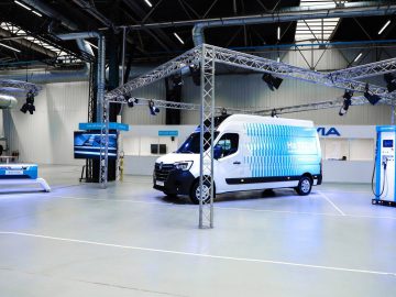 Elektrische bestelwagen tentoongesteld in een moderne industriële faciliteit met als thema H2-waterstoftechnologie. De opstelling omvat informatiepanelen, een laadstation en bovenverlichting.