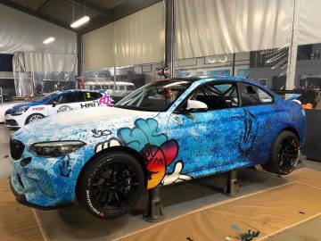 Een felgekleurde blauw-witte BMW M2 CS-raceauto met een kleurrijk graffitikunstwerk-ontwerp wordt in een garage op krikken getild. Op de achtergrond is een andere raceauto zichtbaar, die het talent van Nederlands grootste graffitibedrijf laat zien.