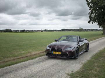 Een bruine BMW M4 Cabrio met een geel kenteken staat geparkeerd op een smalle landweg naast een grasveld onder een bewolkte hemel.