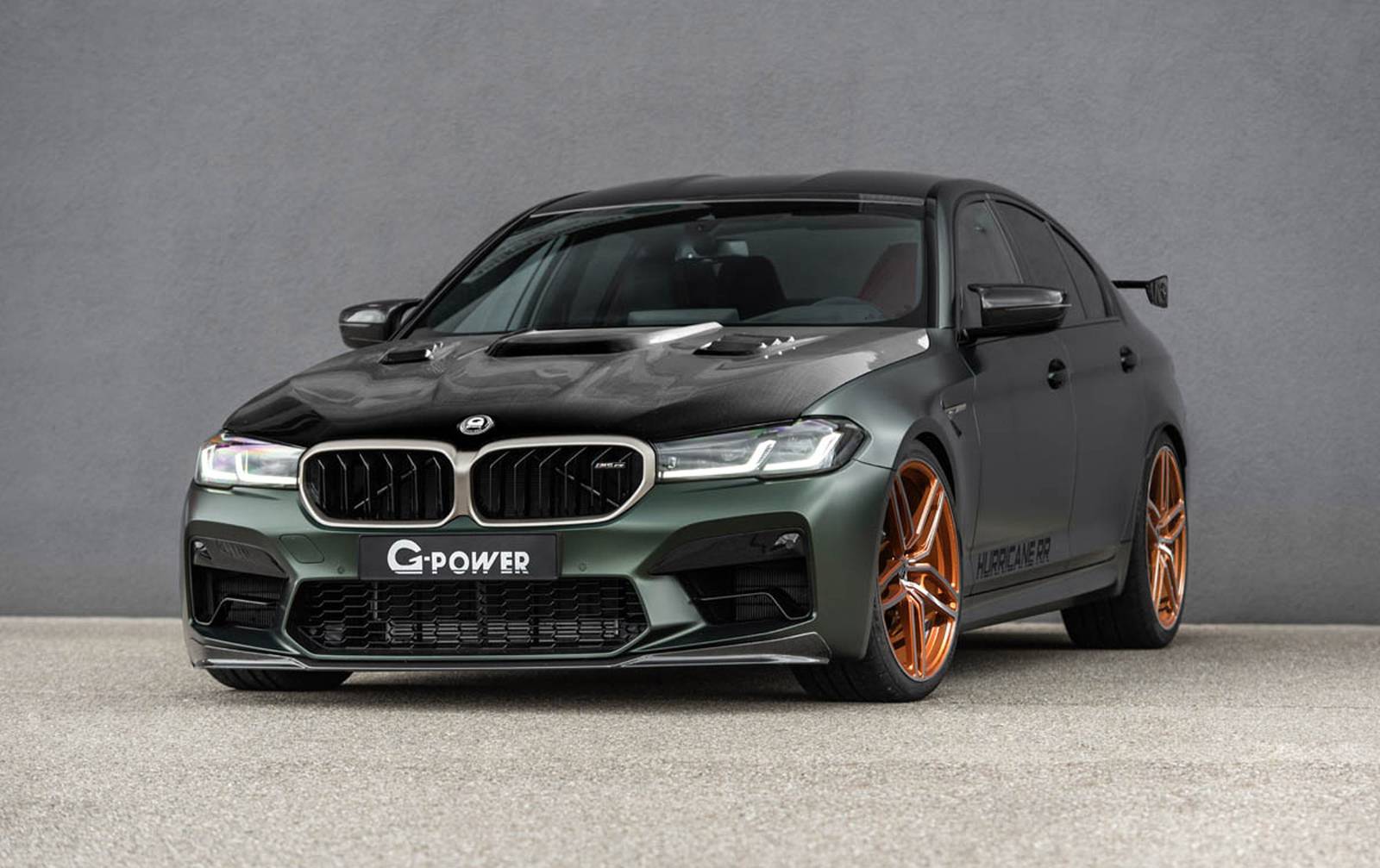 Een aangepaste BMW sedan met een matgroen en zwart exterieur, bronskleurige wielen en een G-Power-badge op de grille, geparkeerd op een betonnen oppervlak tegen een grijze muur.