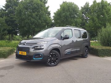 Een grijze Citroën e-Berlingo met zwarte wielen staat geparkeerd op een weg, omgeven door groene bomen en struiken. Het voertuig heeft een Nederlands kenteken met de tekst "P-389-KS.