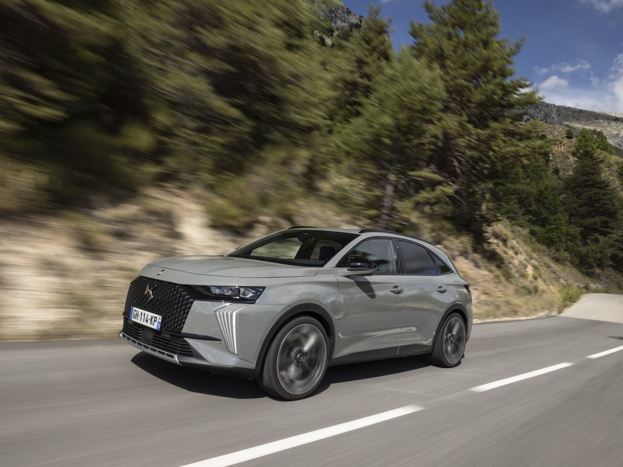 Een slanke zilveren DS 7 SUV rijdt snel over een kronkelige bergweg, omgeven door bomen en rotsachtig terrein onder een gedeeltelijk bewolkte hemel.