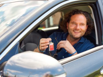 Een man met krullend haar zit glimlachend in een auto en houdt een rode autosleutelhanger van Avis Autoverhuur omhoog.