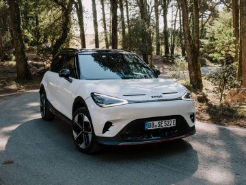 Wit Smart #1 elektrisch voertuig geparkeerd op een bosrijke weg met zonlicht dat door de bomen filtert. Op het kenteken staat "BB SE 522E".