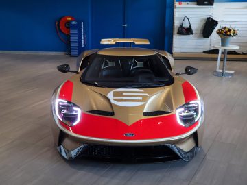 Binnen wordt een gouden en rode Ford GT Holman Woody Heritage Edition met een nummer "6" op de motorkap tentoongesteld. De koplampen van de auto zijn aan en op de achtergrond zijn diverse autoartikelen zichtbaar.