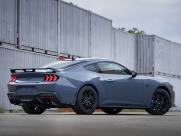 Een blauwgrijze sportwagen, de Ford presenteert gloednieuwe Mustang, staat geparkeerd op een betonnen ondergrond, met zeecontainers op de achtergrond.