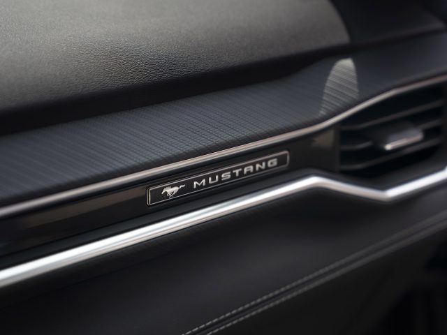 Close-up van het interieur van een auto met het dashboard met het "Mustang"-embleem erop. Het ontwerp toont gestructureerde oppervlakken en een minimalistische stijl, waardoor de gloednieuwe Mustang van Ford wordt benadrukt.