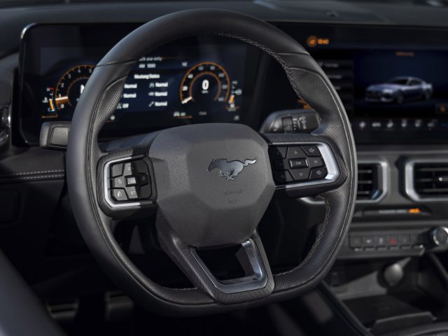 Close-up van het stuur van een moderne auto met een Mustang-logo, met digitale dashboarddisplays en verschillende bedieningsknoppen zichtbaar op de achtergrond. Ford presenteert de gloednieuwe Mustang, met baanbrekende technologie en een strak design.