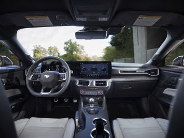 Binnenaanzicht van Ford presenteert gloednieuwe Mustang met een strak dashboard met digitale displays, een stuur met logo en een middenconsole met verschillende bedieningselementen. Lichtgekleurde stoelen zijn zichtbaar.