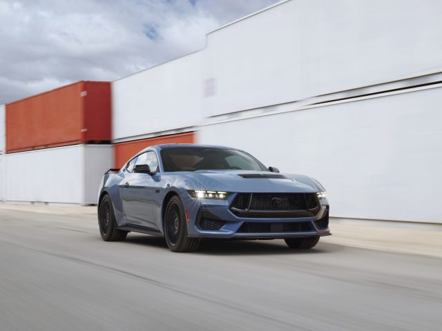 Een strakke blauwe sportwagen rijdt langs zeecontainers op een gladde, open weg onder een bewolkte hemel terwijl Ford een gloednieuwe Mustang presenteert.