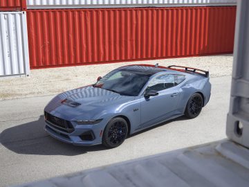 Een grijze sportwagen, die doet denken aan Ford presenteert gloednieuwe Mustang, staat geparkeerd op een verhard oppervlak naast rood-witte zeecontainers. Het ontwerp van de auto is modern met strakke lijnen en een achterspoiler. Er vallen schaduwen op de weg.