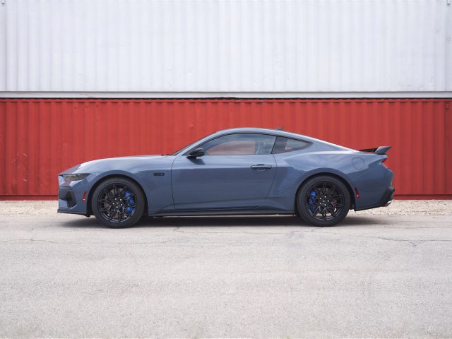 Een blauwe sportwagen met zwarte wielen, geïdentificeerd als de Ford presenteert gloednieuwe Mustang, staat geparkeerd op een betonnen ondergrond voor gestapelde rood-witte zeecontainers.