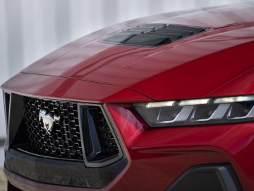 Close-up van de voorkant van een rode sportwagen met een zwarte grille met het embleem van een steigerend paard, hoekige LED-koplampen en een ventilatieopening in de motorkap. Ford presenteert gloednieuwe De Mustang belichaamt de geest van snelheid en design tot in elk detail.