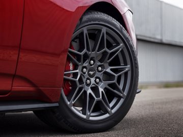 Close-up van een zwarte lichtmetalen velg op een rode auto, met gedetailleerd bandenprofiel en rode remklauw met het Mustang-logo in het midden. In deze verbluffende opname presenteert Ford de gloednieuwe Mustang tegen een achtergrond van een weg en een metalen gebouw.