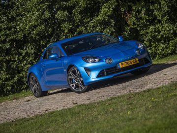 Een blauwe Alpine A110 sportwagen met Nederlands kenteken "P-765-ZB" staat geparkeerd op een stenen pad, omgeven door groen gebladerte.